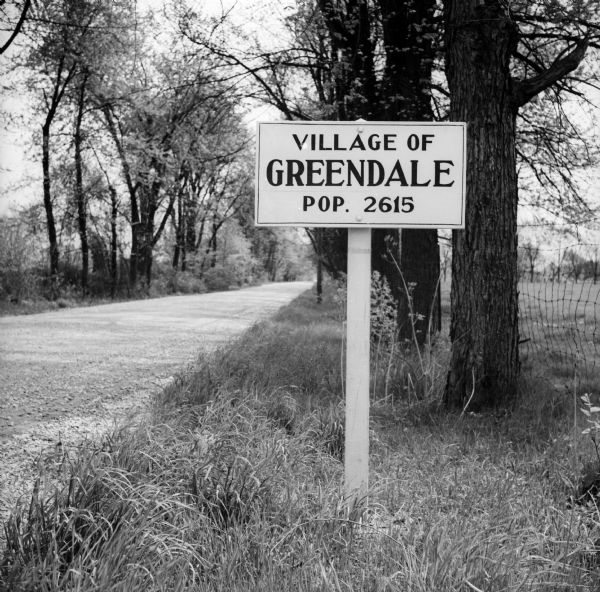 A painted wooden sign on the side of a tree-lined road that reads "Village of Greendale / Pop. 2615."