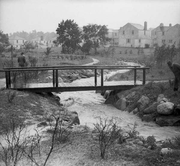 Two men are near a creek, swollen with rainwater, running through Center Park. A footbridge crosses the creek which has large stones along its banks. Trees and other plants are along a footpath which follows the shoreline, and houses are visible in the background.