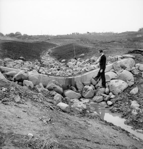 A man stands on rocks that will dam Dale Creek. This picture was likely taken for a study of the water drainage system in early Greendale.