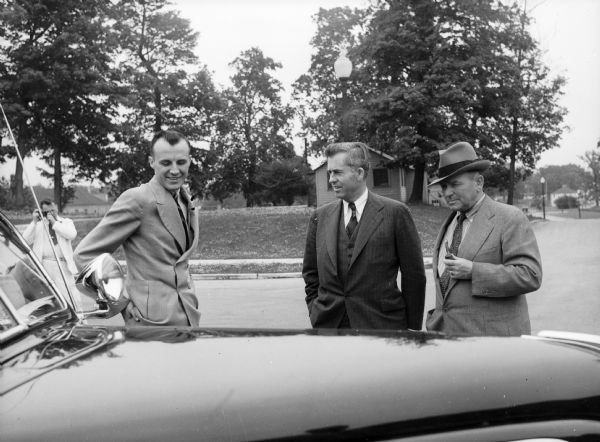 Henry Wallace, between two men, talking outdoors near an automobile. Henry Wallace, then the United States Secretary of Agriculture, visited and toured Greendale. A man in the background is taking a photograph with a camera.