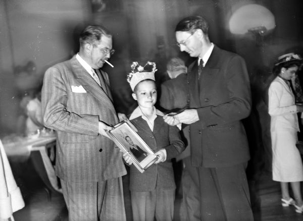 A young boy wearing a crown is presented with a framed portrait of himself by two men, one of whom is smoking a cigarette. The man on the right is holding something in his hand that they are looking at. All three are dressed up and people are milling about in the background near tables in a gymnasium. There is a basketball hoop in the background.