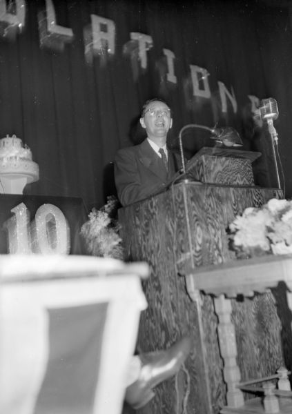 A man stands at a podium speaking into a microphone. There is a cake with the number 10 on it, and letters on the curtain behind him spell out "...gratulations."