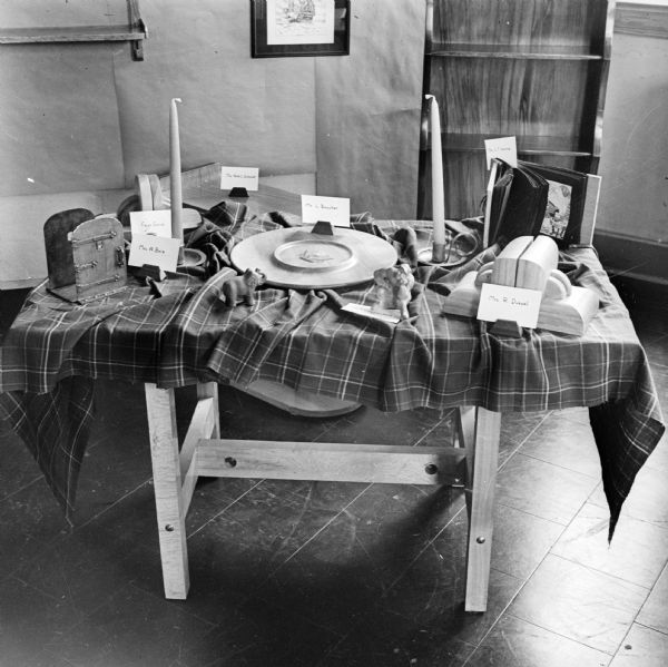 A table spread with crafts at a hobby show with cards identifying the artist. The objects on display include two sets of bookends, one made by "Faye Gore" and the other by "Mrs. R. Duewel," Candles made by "Mrs. A. Boie" and a wooden plate made by "Mr. L. Boucher." There is also a carved elephant, thinking man, and a handmade book, but the names of the creators of those works are obscured.