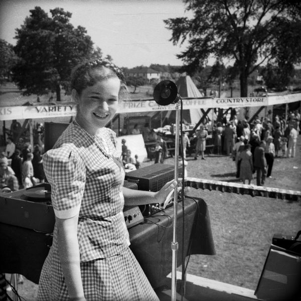 A smiling young girl stands on a raised platform in front of a microphone being used for the Civilian Defense Rally. A record player and other electronic devices sit on a table behind the girl and booths are visible behind the raised platform.