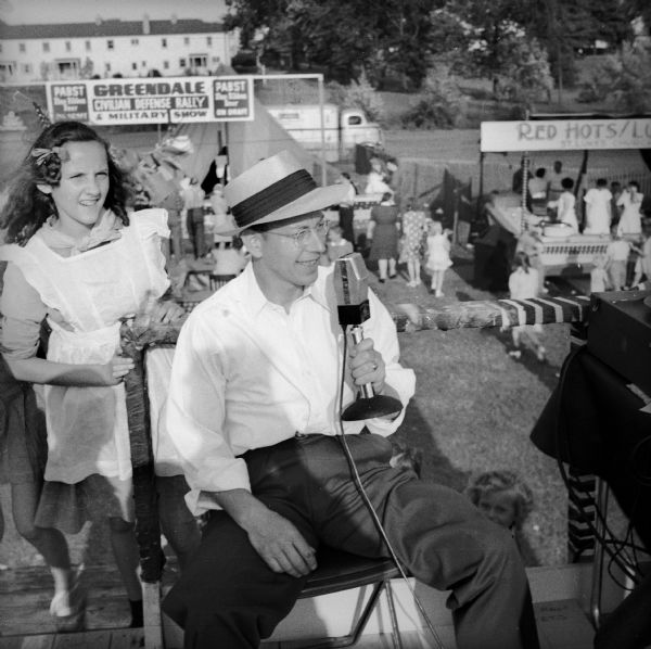 A man wearing a hat and eyeglasses on a raised platform sits holding a microphone. There is a girl standing behind him, and children and adults are around booths visible in the background. One of the booths has a sign for "Greendale Civilian Defense Rally & Military Show." The sign also notes "Pabst Blue Ribbon Beer On Tap."