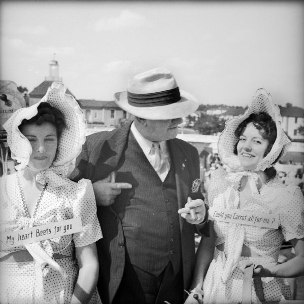A man smoking a cigar and wearing a hat stands between two women wearing bonnets and matching dresses with signs around their necks that read "My Heart Beets For You" and "Could You Carrot All For Me?" The three stand on a raised platform at a Civilian Defense Rally.