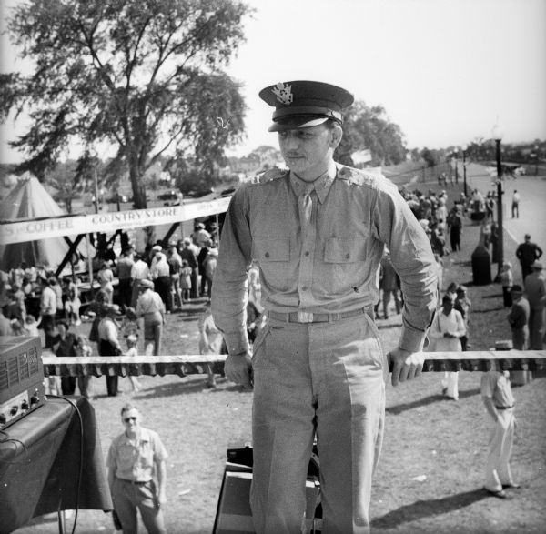 A uniformed member of the US armed forces leans against the rail of a raised platform at a Civilian Defense Rally. The man is wearing a hat and has two bars on his shoulders. People are milling about on the ground behind him near booths, and a street with street lamps is on the right.