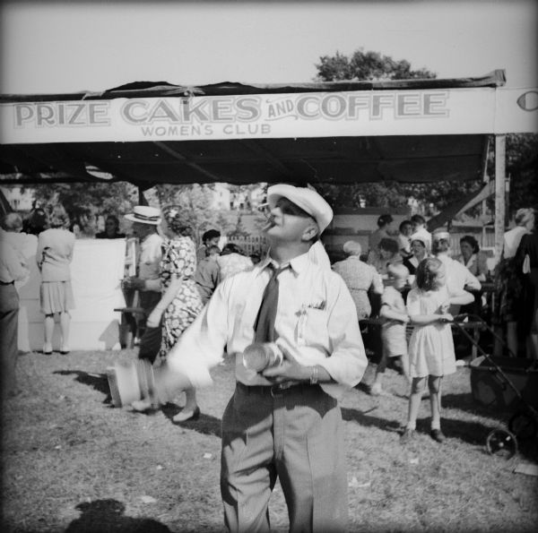 A man smoking a cigarette and wearing a hat is juggling tin cans in front of the "Prize Cakes and Coffee" booth at a Civilian Defense Rally.