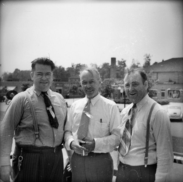 Three men standing next to each other wearing ties on a raised platform at a Civilian Defense Rally. They each have a ribbon on their ties that says: "Committee."