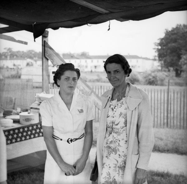 Two women wearing dresses working one of the booths at a Civilian Defense Rally. A table is visible in the background with food on it.