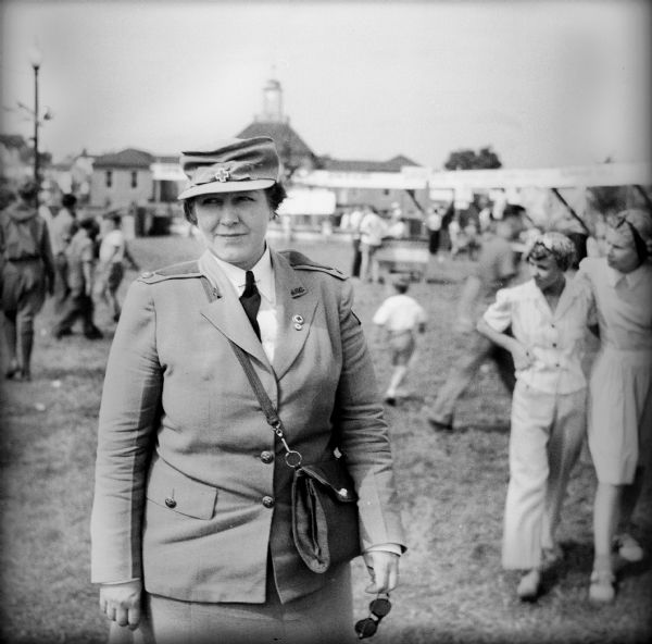 A woman wearing a red cross uniform at a Civilian Defense Rally. Her hat has the red cross logo and she is wearing a red cross lapel pin. The village hall is visible in the background, as are stalls and crowds of people.