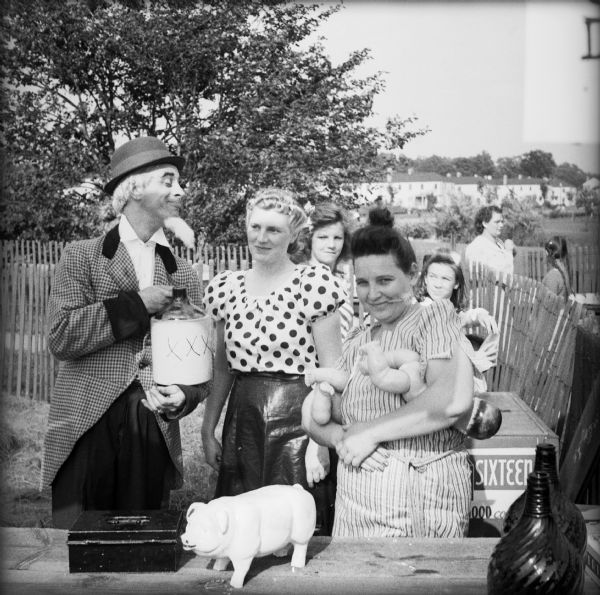 Three people in costume behind a table. There is a man wearing a false beard a hat, holding a jug with three x's on it, a woman wearing a polka-dot blouse and black leather skirt, and a woman wearing a striped dress who is holding two baby dolls. The table has a lock box and a piggy bank on it. The three, as well as others, would perform specific vaudeville stunts as a means of raising money for the war effort.