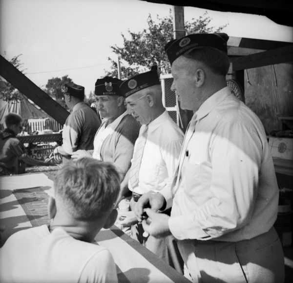 Men from the armed forces working a game booth at a Civilian Defense Rally. Their hats are embroidered with the text "Greendale Wisconsin."