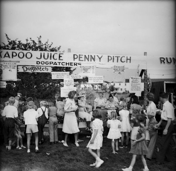 The Dogpathers booth, surrounded by a crowd of children. Text on the banners and other signs read: "Kickapoo Juice Penny Pitch" "Greendale Dogpathers Cause Hitler's Suicide," "Minding Mizzable Kids 10c, Unmizzable 05c, Wet Ones 15c," "Kick Me In The Teeth For 05c". "My Motto I'll Do Anything For A Price," "Wooing Fat Girls 35c, Cross Eyed 25c, Skinny 15c, Ugly 10c, Mizzable 05c," "Have Your Neighbor Bash Me In The Baby For .05c .25c," "Kickapoo Joy Juice Makins: Skunk Innards, Kerosene, Catfih Haids, Corn Squeezins, Dale Creek Water." The Dogpatchers would perform specific vaudeville stunts as a means of raising money for the war effort.