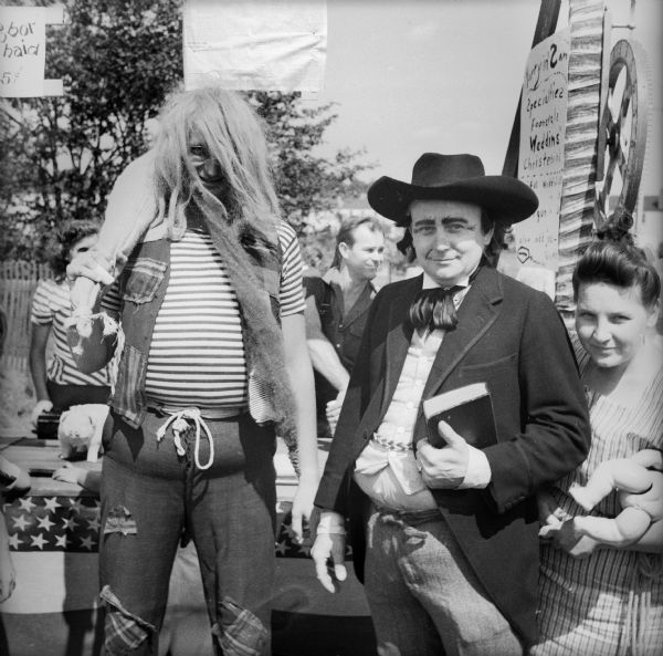 A man with a gunny sack swung over his shoulder is dressed as a hillbilly with a wig and a fake beard. Next to him stands a man dressed as a preacher, and a woman holding two baby dolls. The three, as well as others, would perform specific vaudeville stunts as a means of raising money for the war effort. A sign in the background reads "Marryin Sam Specialties, Foonerals, Weddins, Christenins."