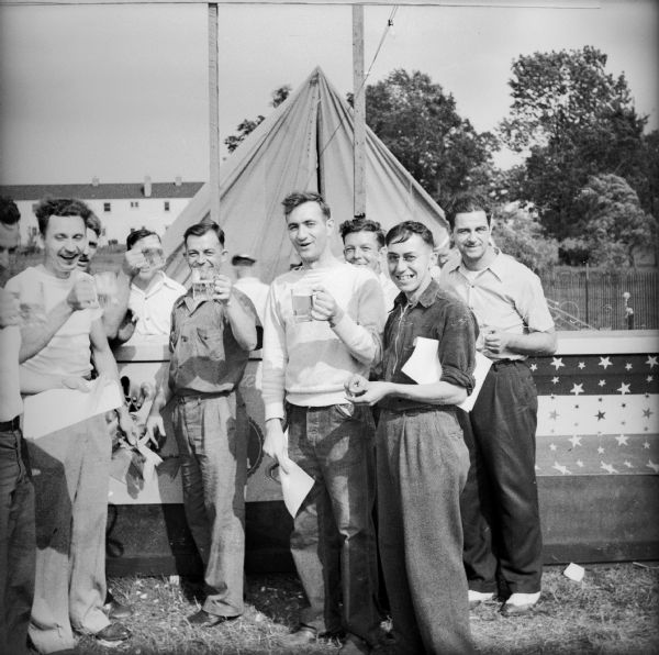 A group of men standing next to an outdoor bar raise mugs of beer at a Civilian Defense Rally. There is a tent and dwellings in the background.