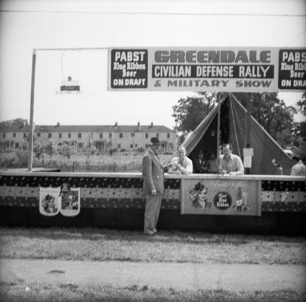 A man orders a beer from the outdoor bar at a Civil Defense Rally. The banners on the booth read "Pabst Blue Ribbon Beer On Draft" and "Greendale Civilian Defense Rally & Military Show." There is a tent in the background.