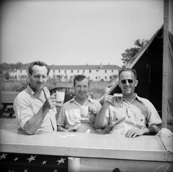 Three men raising mugs of beer from behind the outdoor bar at a Civilian Defense Rally. In the background is a tent.