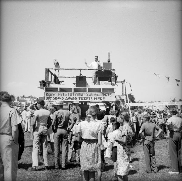 A crowd of people mills around the Civilian Defense Rally ticket booth, which has an announcer situated on the roof. He is speaking into a microphone, addressing the crowd. The sign on the ticket booth reads "Register Here For Free Chance On Attendance Prizes Buy Grand Award Tickets Here."