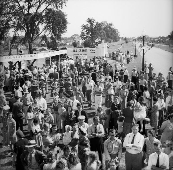 Elevated view from the announcer's booth of crowd of people at a Civilian Defense Rally. The ice cream stand and the "country store" are visible in the background, and in the far distance are houses.
