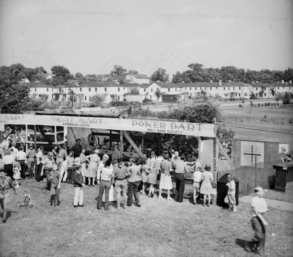 Elevated view of people crowding around booths at a Civilian Defense Rally, "Poker Dart, Holy Name Society," "Meat mart, American Legion," "[obscured]Jap! [obscured]." A sailor in uniform and a Cub Scout are visible in the crowd. In the background are houses, and a barn with a silo.