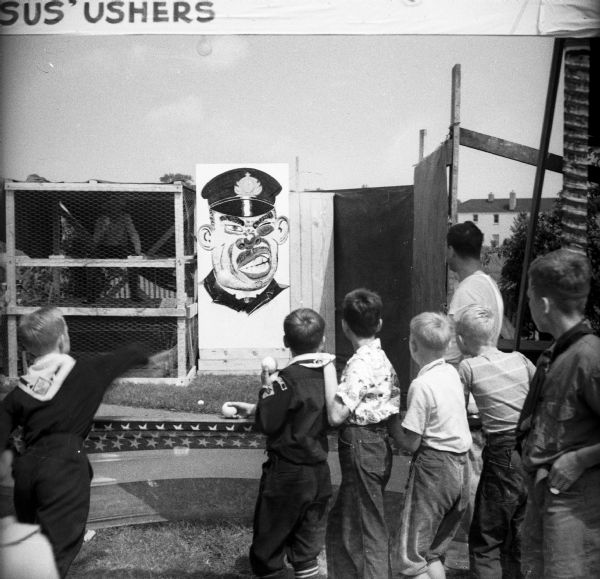 Cub Scouts throw baseballs at a large caricature of a Japanese soldier during a Civilian Defense Rally. A man is sitting in a dunk tank on the left in a box surrounded by chicken wire.
