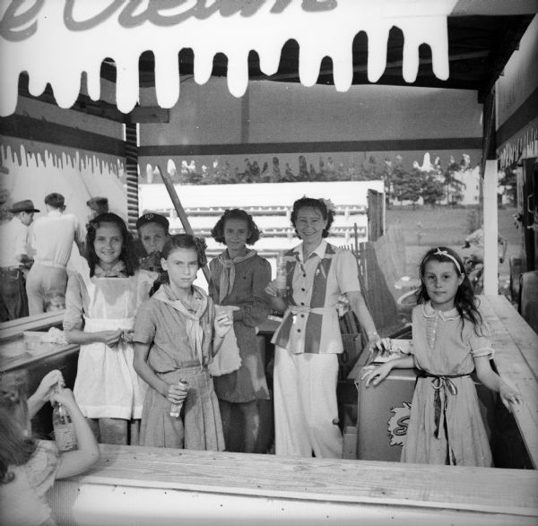 A group of girls working behind the counter at the ice cream booth at the Civilian Defense Rally take a moment to pose for a picture. Two of the girls are wearing girl scout uniforms.