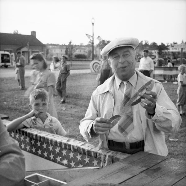 A man wearing a hat and tie holds tickets he just purchased for use at a Civilian Defense Rally. A child stands to his right leaning on the ticket counter. In the background are children and adults near booths.