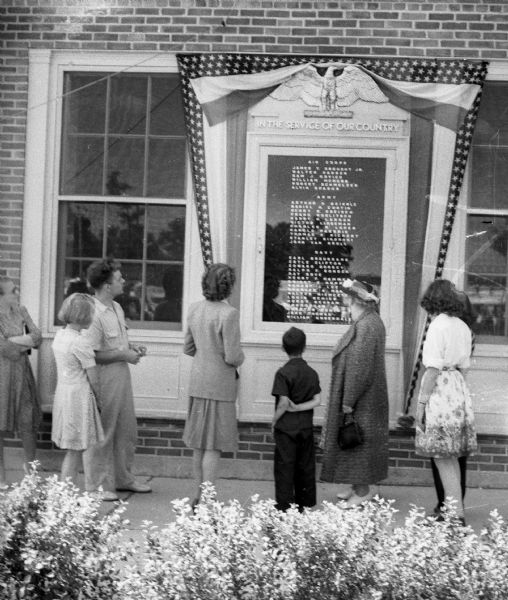 A group of people standing in front of a list posted outside of a building of all the members of the Greendale, Wisconsin, community who are "In The Service Of Our Country" in the various branches of the armed forces. The list is broken down by Air Corps, Army and Navy. 

"Air Corps:

James Y. [obscured] Jr. 
Walter Hansen, 
Sam J. Kovich 
William McNabb 
Robert Schmelzer 
Alvin Parks 

Army: 

Arthur H Beierle 
Daniel H. Curtin 
Henry B. Greisen 
Frank Kuglitsch 
Harry H. Larson 
Voctor Lingeman 
Chrles W. Loper 
Harold Pasbrig 
Thomas E. Rickert 
Stanley Sikorski 
Johnny Van Groll

Navy: 

Earl Broussard 
Estle Clifton 
Edward Dorsey 
Ralph F. Gondek 
Ray Gove 
Henry F. Hammer 
Dwight E. Hoppert 
Robert J. Luzinski 
Charles A. Murdaugh 
Walter Niemczyk 
Louis Schier 
Horst Schram 
John Shaw 
Chester Sowin 
John Thoman 
William Underdale."