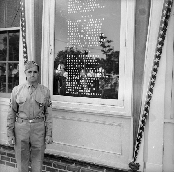A soldier in full uniform stands next to a list of all the members of the Greendale, Wisconsin, community who are enlisted with the armed forces. The list is broken down by Air Corps, Army and Navy.

"Air Corps:

James Y. [obscured] Jr. 
Walter Hansen, 
Sam J. Kovich 
William McNabb 
Robert Schmelzer 
Alvin Parks 

Army: 

Arthur H Beierle 
Daniel H. Curtin 
Henry B. Greisen 
Frank Kuglitsch 
Harry H. Larson 
Voctor Lingeman 
Chrles W. Loper 
Harold Pasbrig 
Thomas E. Rickert 
Stanley Sikorski 
Johnny Van Groll

Navy: 

Earl Broussard 
Estle Clifton 
Edward Dorsey 
Ralph F. Gondek 
Ray Gove 
Henry F. Hammer 
Dwight E. Hoppert 
Robert J. Luzinski 
Charles A. Murdaugh 
Walter Niemczyk 
Louis Schier 
Horst Schram 
John Shaw 
Chester Sowin 
John Thoman 
William Underdale."