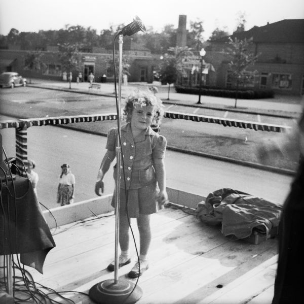 A young girl stands behind a microphone on the announcers platform at a Civilian Defense Rally. In the background are a road and commercial buildings.