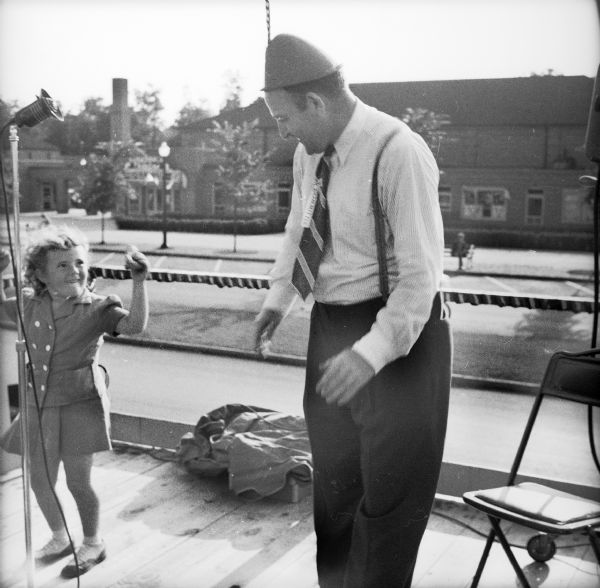 A young girl dances with an older man wearing a cone-shaped hat and a tie on the announcers platform at a Civilian Defense Rally. To the far left a microphone is visible, and a road and commercial buildings are in the background.