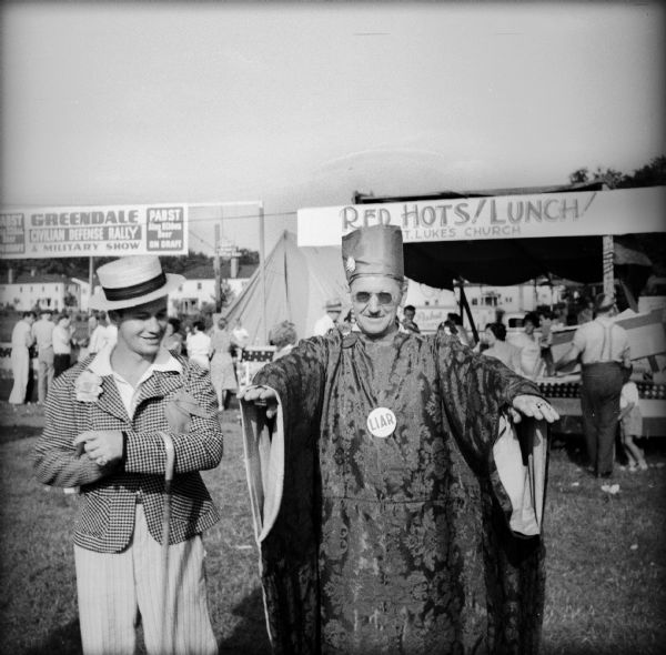 Two men wearing costumes as a part of a Civilian Defense Rally. The man on the left is wearing a straw hat, seersucker jacket, corsage, with a cane hanging from his left arm. The gentleman on the right is holding out his arms and wearing a turban-like hat, a paisley patterned robe, and a button with the word "liar." In the background the outdoor bar is visible, as is the "Red Hots! Lunch!" booth which is being run by St. Luke's Church.