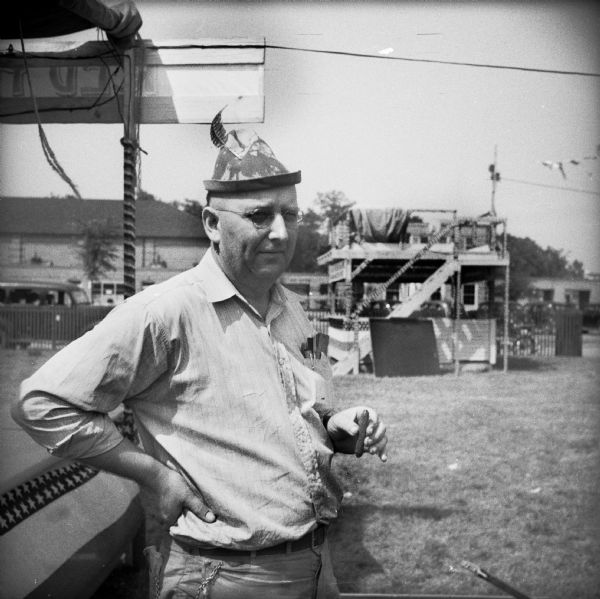 A man near a booth wearing a hat with a feather smokes a cigar while setting up for a Civilian Defense Rally. The announcers platform is in the background.