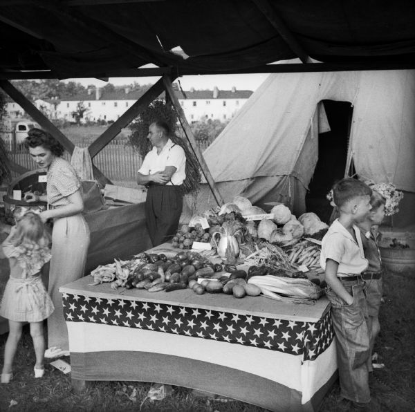Adults and children stand around a vegetable stand under a covered booth at a Civilian Defense Rally. Behind the booth is a tent, and in the far background is housing.