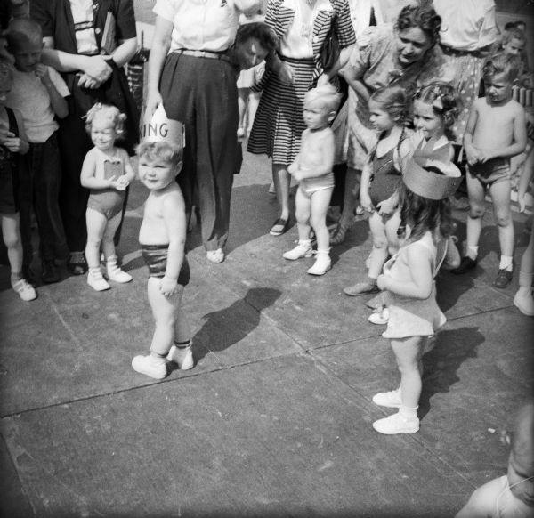 Young boys and girls participate in a "Childrens Bathing Beauty Contest" at a Civilian Defense Rally. Women are standing around the children. The winners, 2-year-old children Tom Glyn and Donna Marie Mason, king and queen respectively, are wearing paper crowns.