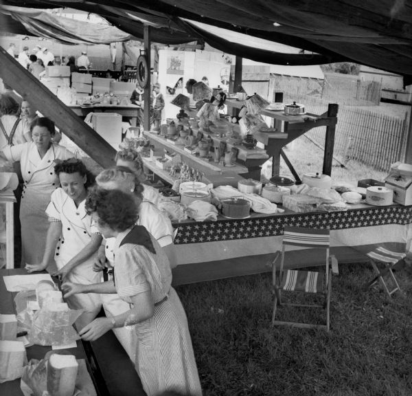 Side view of a group of women at a Civilian Defense Rally standing behind a counter with loaves of bread. Behind them is a table of covered cake pans and other baked goods. More booths can be seen in the background.