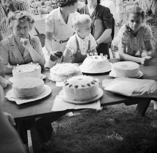 A woman and children sit around a picnic table with a variety of cakes displayed.