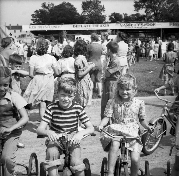 A group of children pose on tricycles at a Civilian Defense Rally. Booths and crowds are visible in the background, including a game booth where you can "Deflate The Axis" which is being run by the Cub Scouts.
