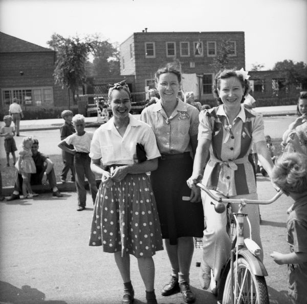 Three women at a Civilian Defense Rally, one of whom is sitting on a bicycle. Children and adults are standing in the street and on the curb in the background.
