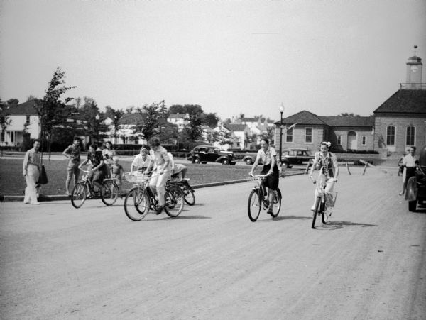 Girls riding bicycles at a Civilian Defense Rally. The Greendale Village Hall with clock tower is visible in the background, and the street has been blocked off by wood barriers.