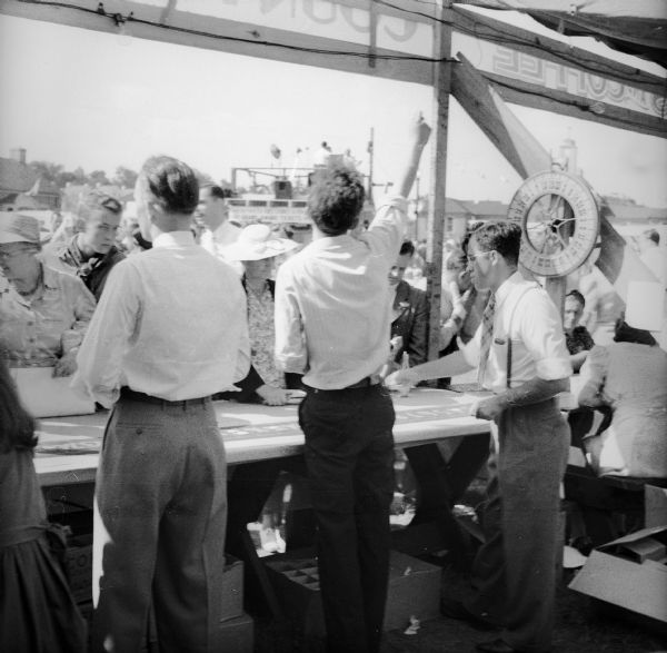 Rear view of men working the "Country Store" booth at a Civilian Defense Rally. A "wheel of fortune" device is on the right and a crowd and an elevated announcers platform are in the background.