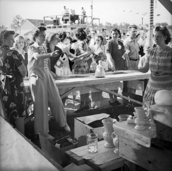 Women selling ceramics behind a booth at a Civilian Defense Rally. The elevated announcers booth is visible in the background.