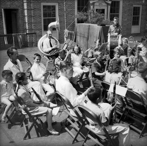 A group of school-age children with a several adults sitting outdoors with band instruments at a Civilian Defense Rally.