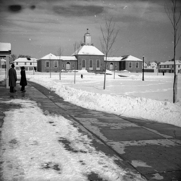 Two adults looking at the Greendale Village Hall with clock tower across an expanse of snow.