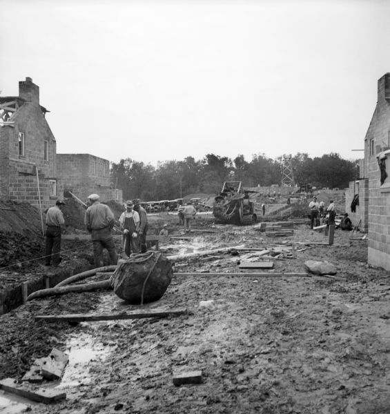 Men work to level the ground for foundations to be poured for homes. Exposed brick walls of partially constructed homes are on the left and right, and a ditch digging machine.