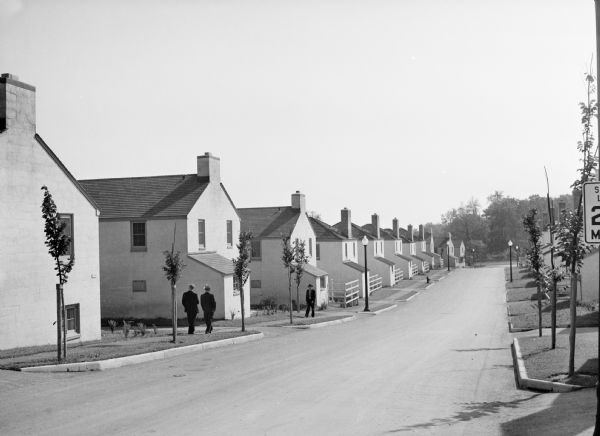 View up residential street with newly constructed houses, young trees and lampposts. Men in suits and hats walk along the sidewalk. A speed limit sign is on the right.