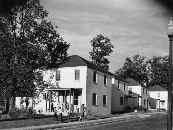 View from road of a group of seven children playing in front of a house and on the porch, while a woman holding a bag of groceries walks past on the sidewalk. A street lamp is visible along the right side of the image.