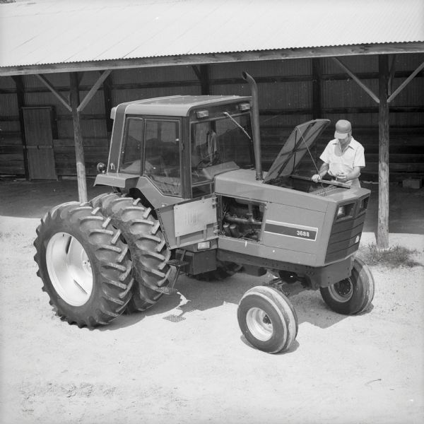 Man with International Harvester 3688 tractor with maintenance panels open near shed roof overhang.