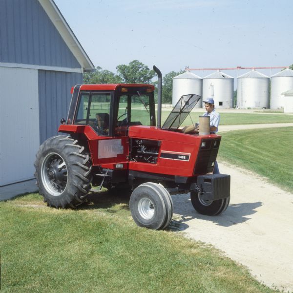 Color photograph of a man working on an International Harvester 5488 tractor with maintenance doors open on farmstead. There are silos in the background.
